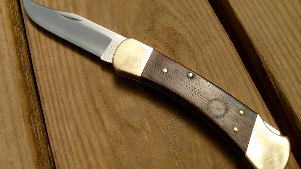 Buck-style lockback folding knife with wood handle and brass bolsters on a wooden deck. Representative photo, not the exact product.