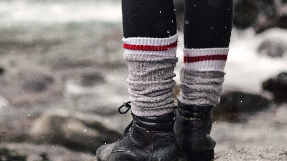 Heavy red-striped wool hiking socks above black boots on a rock by a stream. Representative photo, not the exact product.