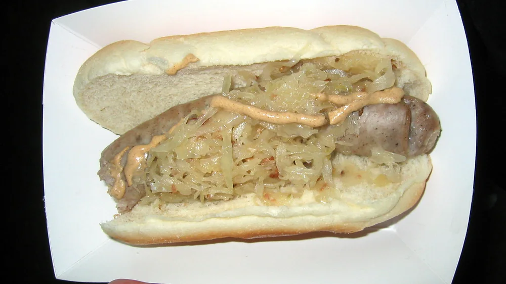 A cooked Usinger's bratwurst with sauerkraut on a roll in a paper tray. Representative photo of the product, not a gift box.