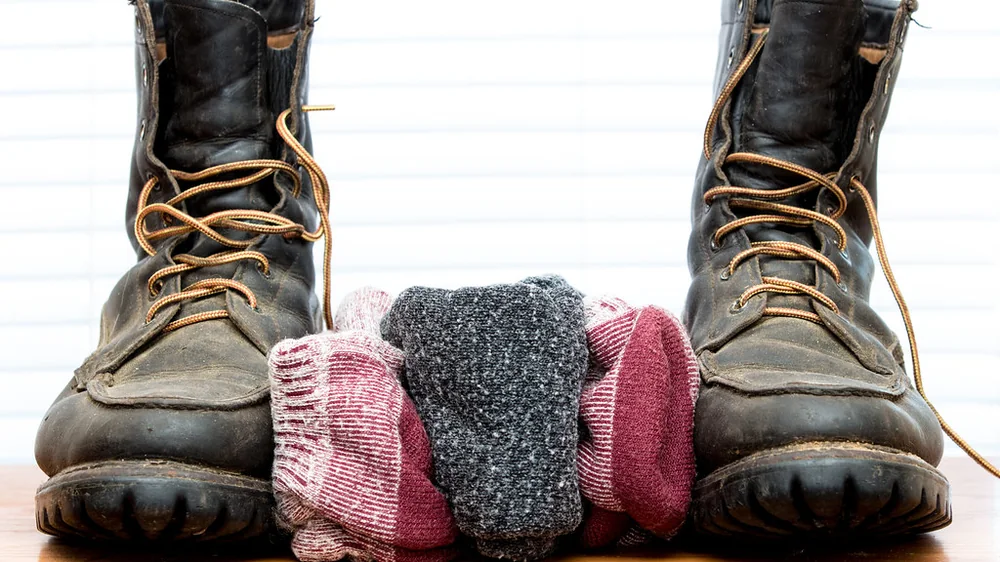 Folded wool socks in several colors between a pair of old leather work boots on a shelf. Representative photo, not the exact product.